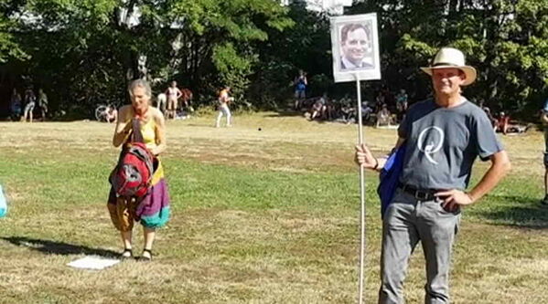 Sie trägt ein dunkles Ein Mann im T‑Shirt mit einem großen „Q“ auf der Vorderseite und hält ein Plakat mit dem Porträt eines Mannes darauf.