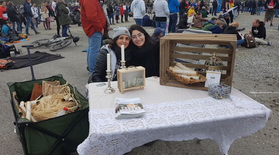 Zwei junge Damen an einem Tisch mit Kerzen, Flyern und Schriftrollen und einer Kiste mit einer Krone sitzend an einem Tisch