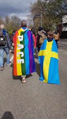 Regenbogen‑Friedensflagge mit dem Schriftzug ‚PACE‘ und eine schwedische Nationalflagge als Umhang