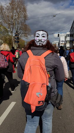 Person mit Maske am hinterkopf, Rücksack darunter -- knallorange