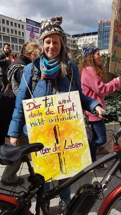 Fahrradfahrerin mit Plakat mit Kreuz vor kräftig orangem Hintergrund.