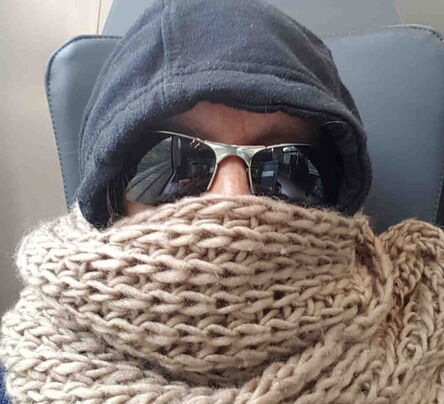 Person mit grobmaschigem hellbrauchen Schal um Mund und Nase, eine Sonnenbrille und eine Mütze