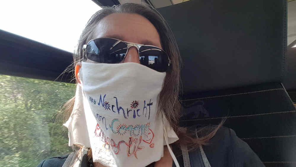 Frau mit Sonnenbrille im Zug - ein weißes Leinentuch um Mund und nase, mit Aufschrift.